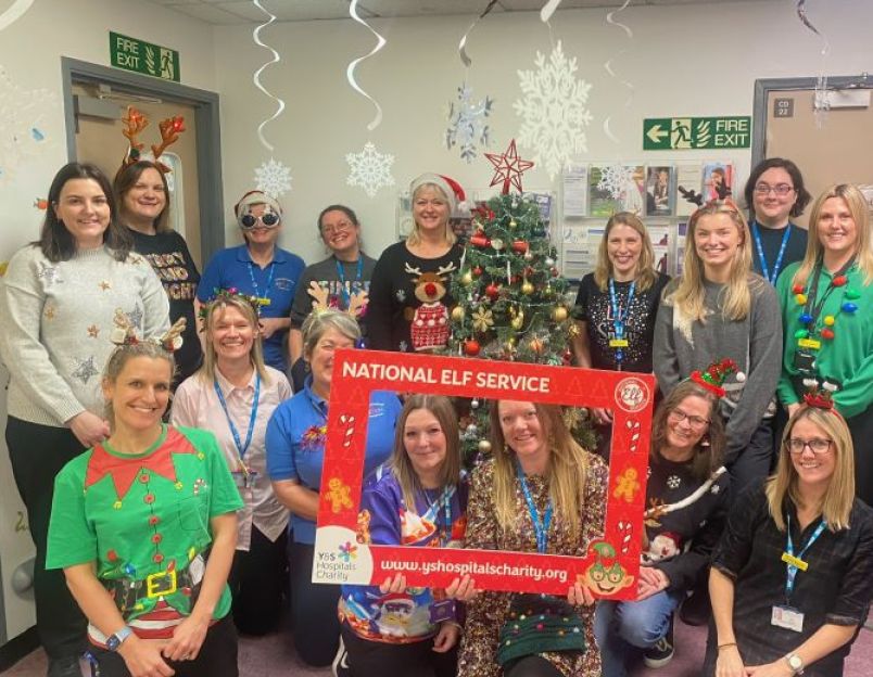 A large group of staff wearing Christmas jumpers, smiling, standing by a Christmas Tree