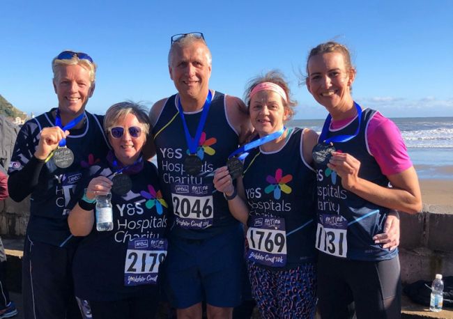 Five supporters show their medals after completing a 10k race. They are standing by the beach 